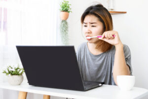 workaholic Asian woman busy with working hard on computer laptop another hand brushing teeth sitting at desk